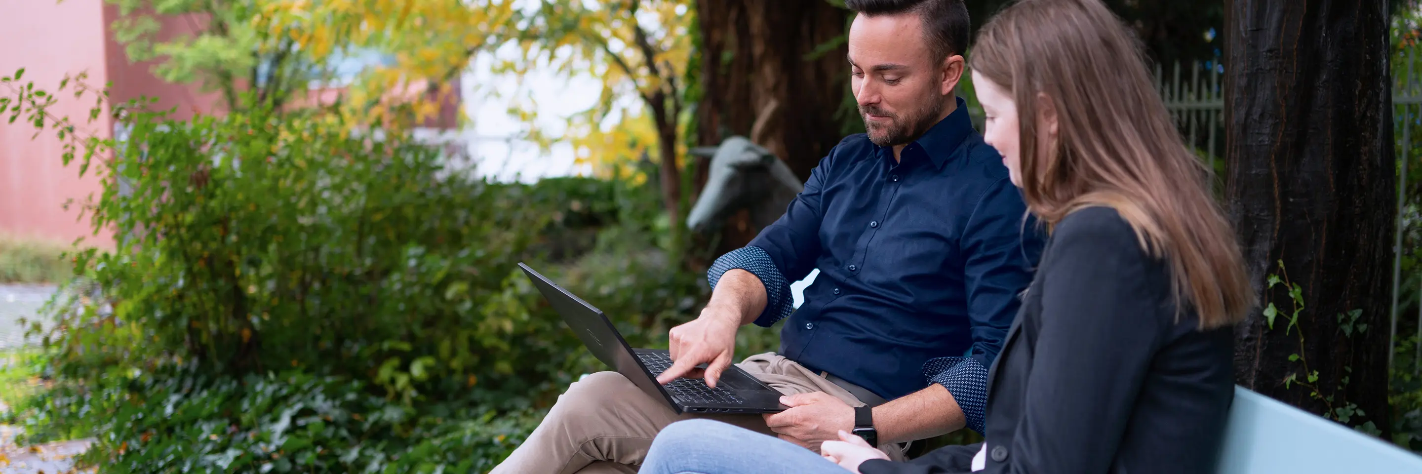 Zwei Personen sitzen draußen auf einer Bank und schauen gemeinsam auf einen Laptop